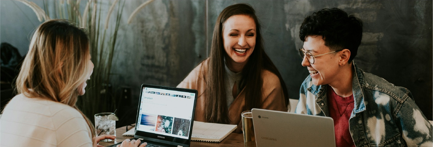 Three people sit at a table, laughing with each other as they work on laptops