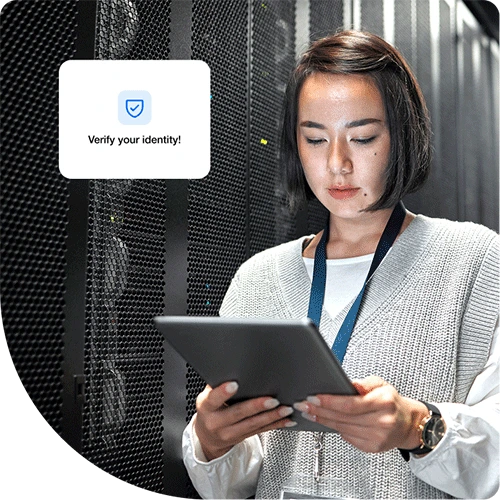 A woman with short dark hair is standing in a server room, holding and working on a Duo trusted device. A digital message box on the left side of the image displays a shield icon and the text 